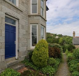 An exterior view of a stone house with a blue door and garden path surrounded by plants and bushes at Lookout in Mousehole