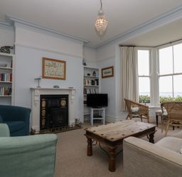 A living room with a fireplace bookshelves armchairs a wooden coffee table a sofa and a bay window with wicker chairs at Lookout in Mousehole