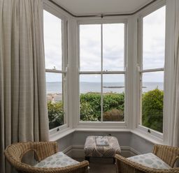 A seating area with two wicker chairs and a patterned ottoman by a bay window overlooking greenery and the sea at Lookout in Mousehole