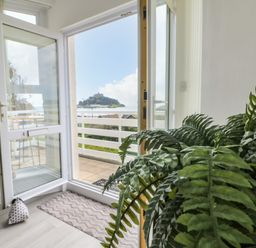 An open glass door leading to a balcony with a view of a hill and sea with a large green plant inside at Sea Breeze in Marazion