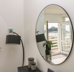 A round wall mirror reflecting an open door with a view of a castle across water beside a floor lamp plant and small containers at Sea Breeze in Marazion