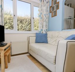 A small living room with a white leather sofa and blue cushions beside a window and a wooden side table with a television at Respryn Mevagissey
