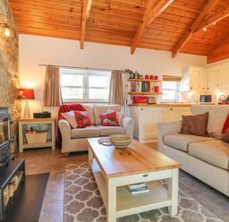 A living room with two sofas, a coffee table, a wood stove, and a kitchen area at Pond Cottage in Stithians