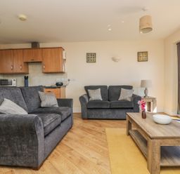 A living room with two grey sofas a wooden coffee table with flowers and a bowl and a kitchen area at Surfside in Marazion