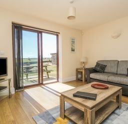 A living room with a sofa a wooden coffee table a television on a glass stand and sliding glass doors leading outside at Seashells in Marazion