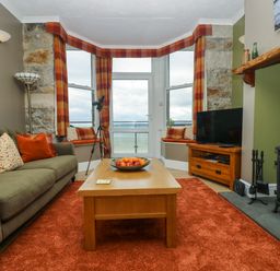 A living room with a green sofa red cushions a wooden coffee table with fruit a large window with plaid curtains a television and a wood burning stove at Mount View in Marazion