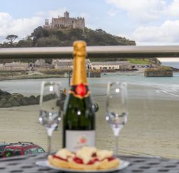 A bottle of champagne two empty glasses and a plate with food on a table overlooking a sandy beach and a castle on a hill at Mount View in Marazion