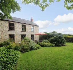 A stone house with multiple windows a garden with bushes green grass and a patio table with an orange umbrella at Chenalls Cottage in Helston