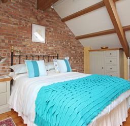 A bedroom with a bed covered with a turquoise blanket and pillows against a brick wall with wooden furniture and a standing mirror at Summer House in Helston