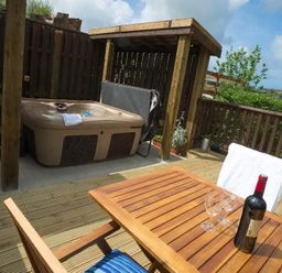 An outdoor deck area with a hot tub under a wooden pergola a wooden table with wine bottle and glasses and wooden chairs at Godrevy Cottage in Hayle