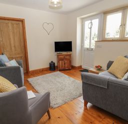 A living room with grey sofas and armchair around a rug a wooden door and a small TV on a wooden stand at Honeysuckle Cottage in Helston
