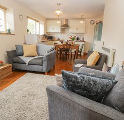 A living room with grey sofas, a wooden dining table and chairs, kitchen cabinets, and wooden floor at Honeysuckle Cottage in Helston