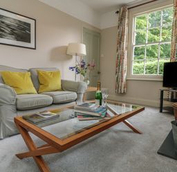 A living room with a sofa two yellow cushions a glass coffee table with books and a bottle a TV on a stand and a window with patterned curtains at Penwarne Lodge in Mawnan Smith