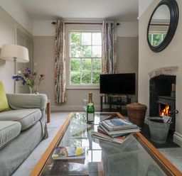 A living room with a sofa a glass coffee table a fireplace a television and a window with curtains at Penwarne Lodge in Mawnan Smith