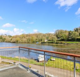 A balcony overlooking a river with a boat on the shore and trees in the background at Carne Mill in Manaccan