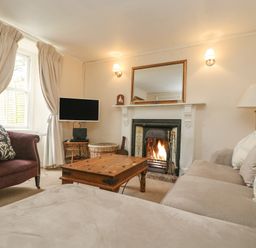 A living room with a beige sofa with cushions a wooden coffee table a purple armchair a fireplace with a fire and a wall-mounted television at Little Chycoose in Truro