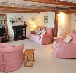 A living room with exposed wooden beams pink upholstered sofa and chairs a fireplace and wooden furniture at Rose Cottage in Manaccan
