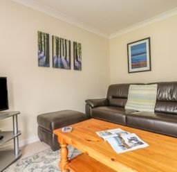 A living room with a brown leather sofa a wooden coffee table a TV on a stand and framed pictures on beige walls at Cuckoo's Cottage in Falmouth