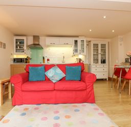 A living room with a red sofa and blue cushions facing a kitchen with white cabinets and a dining area with red chairs at Plum Cottage in Colebrooke near Crediton