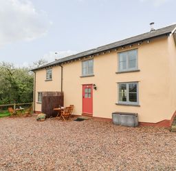 A house with a red door and two sets of wooden tables and chairs on a gravel driveway at Apple Cottage in Colebrooke near Crediton
