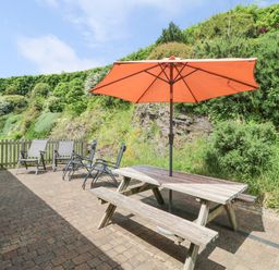 An outdoor patio with a wooden picnic table and orange umbrella surrounded by garden chairs and greenery at Roosters in Lostwithiel