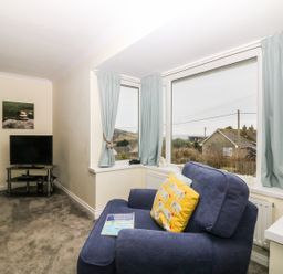 A living room with a blue armchair facing windows with light blue curtains and a television on a black stand at Bethany Bay View in Holywell Bay