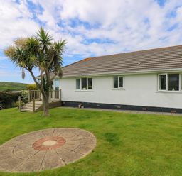 A single story house with a tiled roof and white walls with a small tree and circular paved area in the grassy garden at Bethany Bay View in Holywell Bay
