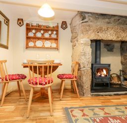 A dining area with a wooden table and three chairs with red cushions near a stone fireplace with a wood-burning stove at Gurnard's Cottage in St Ives
