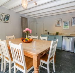 A dining room with a large wooden table surrounded by white chairs a bowl of fruit and flowers on the table blue cabinets with a dishwasher and framed pictures on the wall at Parwin in Tintagel