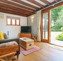 A living room with wooden beams on the ceiling a leather sofa cushions a wooden chest as coffee table a television on a wooden cabinet and glass doors opening to a garden at Oaklands Near Boscastle