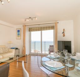 A living room with a glass dining table set for four and a sofa facing a glass coffee table with sea view at Seasiders Ilfracombe