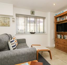 A sitting area with a gray sofa and striped cushions next to a window with blinds and a wooden media cabinet at Seascape in Porthcurno