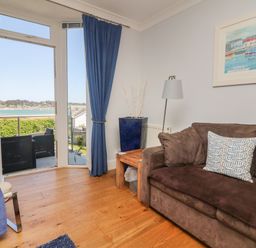 A living room with a brown sofa blue curtains and a door opening to a balcony overlooking water at Tides in Padstow