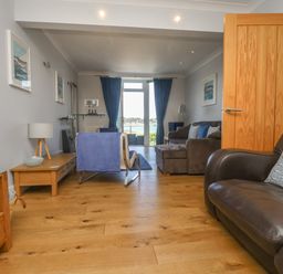 A living room with wooden flooring sofas a chair tables blue curtains and a door with a waterfront view at Tides in Padstow