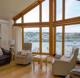 A sitting area with two armchairs a sofa and a wooden table in front of large windows overlooking a marina at Totherside in Kingswear
