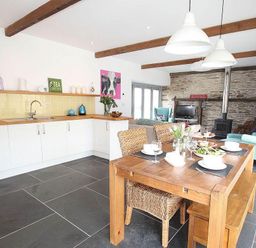 A kitchen and dining area with wooden table and chairs and a living room with teal armchairs and a wood stove at The Old Dairy in Padstow