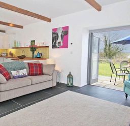 A living area with a beige sofa with red pillows and a blue armchair next to an open door leading to a patio with a blue umbrella at The Old Dairy in Padstow