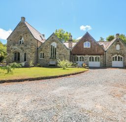 A stone house with multiple gable roofs a gravel driveway and a lawn with plants and trees at Hideaway in Watergate Bay