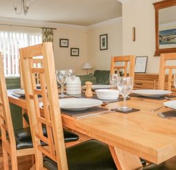 A dining room with a wooden table set with plates glasses and cutlery surrounded by wooden chairs with a green sofa in the background at Fairways in Woolacombe