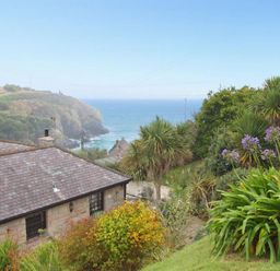 A garden with various plants and a bench overlooking a stone house roof and the ocean at Mengarth in Cadgwith