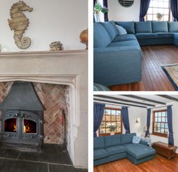 A living room with a stone fireplace with fire a blue sectional sofa wooden floors and windows with blue curtains at Mengarth in Cadgwith