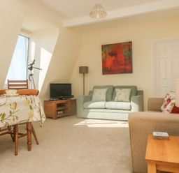A living room with a dining table a green sofa a beige armchair a wooden TV stand a floor lamp and a telescope at Gannet Watch in Coverack