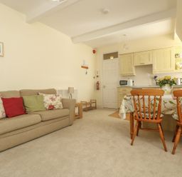 A living room with a beige sofa and colorful cushions a dining table with wooden chairs and a kitchen area at Gannet Watch in Coverack