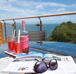 A glass table with two glasses of pink drink a bottle of raspberry lemonade an open magazine and black sunglasses on a balcony with a sea view at Bayview in Brixham