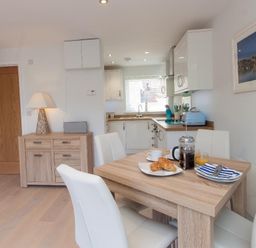 A dining table with breakfast items and white chairs in a kitchen with wood cabinets and a sideboard with a lamp at Bayview in Brixham