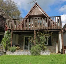 A two-story house with a wooden balcony a grassy lawn and trees at Devine Lodge in Callington