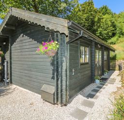 A wooden cabin with hanging flower baskets and a stone pathway surrounded by trees at Pond Cabin in Stithians