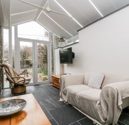 A room with a beige sofa covered with a throw and a cushion a rattan chair a wooden table with a decorative bowl and a wall mounted TV at Forge Cottage in Sennen