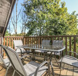 An outdoor wooden deck with a glass table and six chairs surrounded by trees at Valley Lodge 58 in Callington