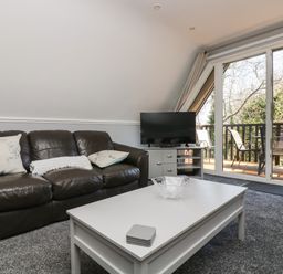 A living room with a brown leather sofa, white coffee table, TV on cabinet, and sliding glass doors to a balcony at Valley Lodge 58 Callington